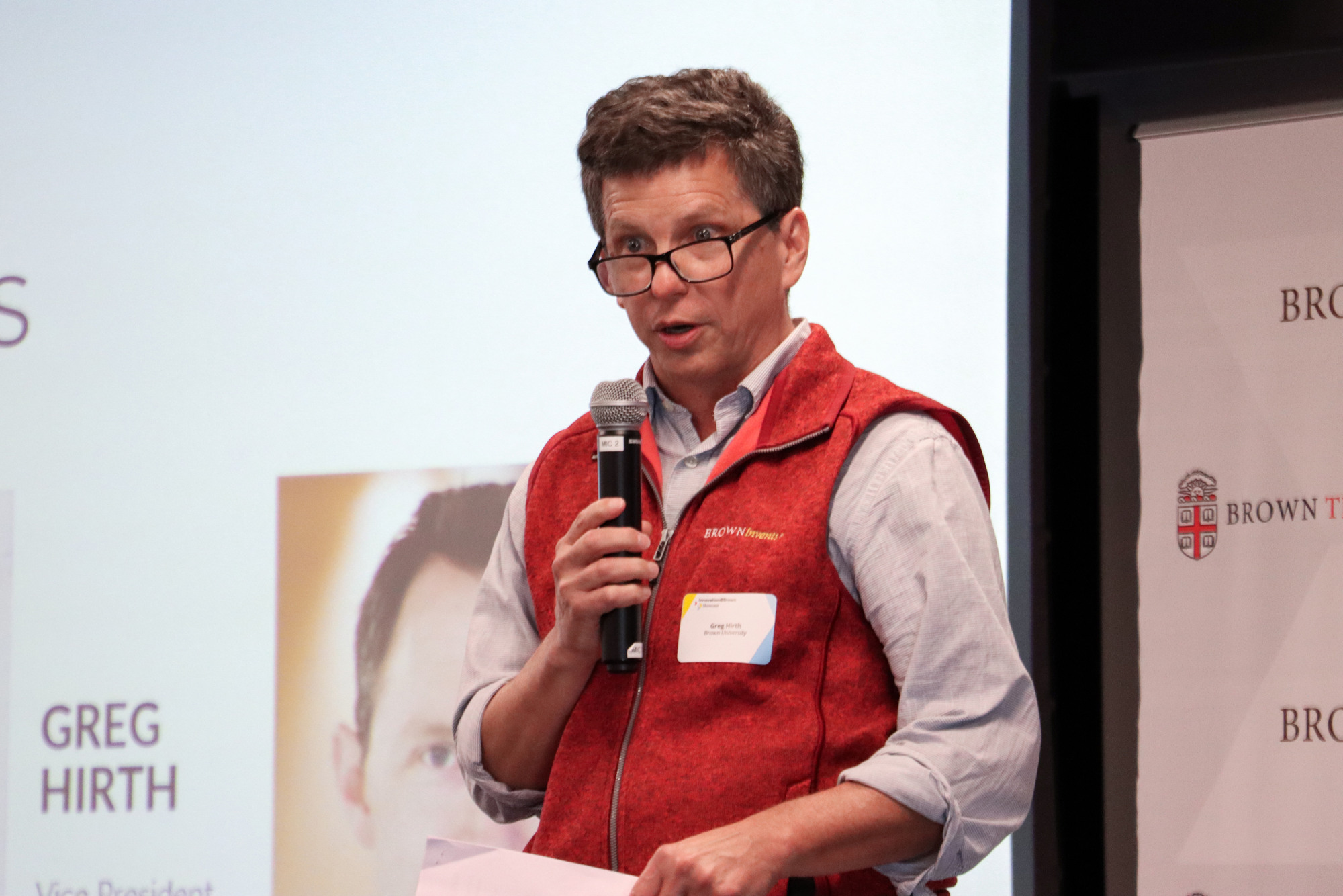 Greg Hirth speaks on stage and holds a microphone to his mouth.